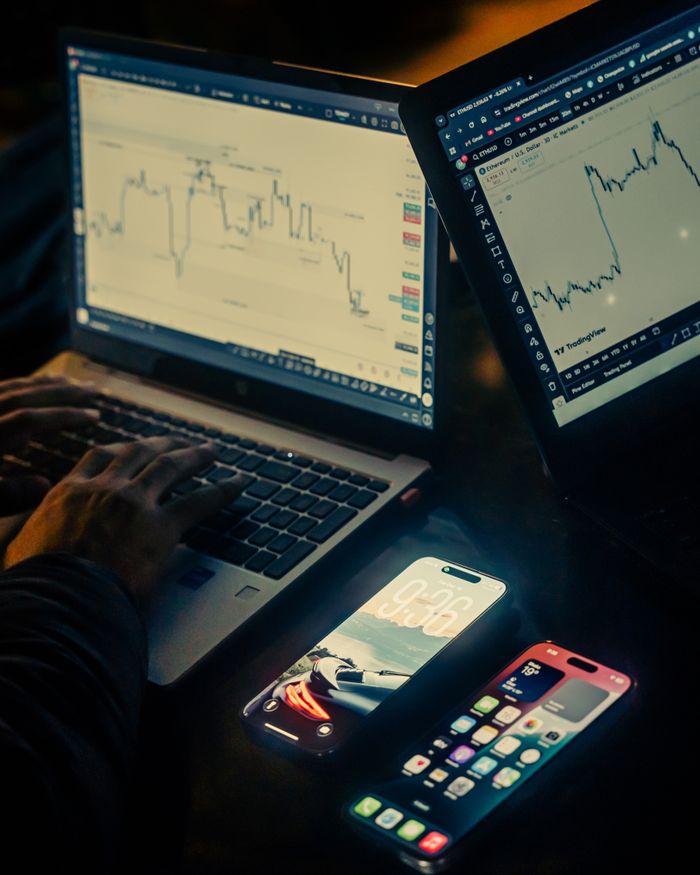 Un espace de travail de trading avec des ordinateurs portables affichant des graphiques et des smartphones, dans une ambiance peu éclairée, à Dhaka.